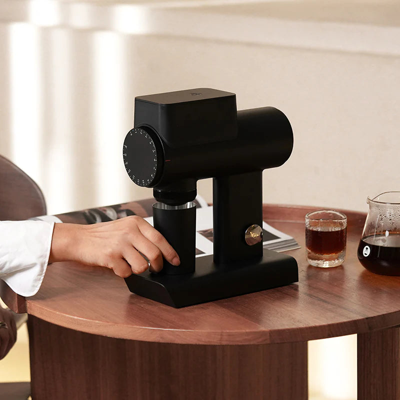 Black coffee grinder on a wooden table with a glass of coffee and a carafe in the background.