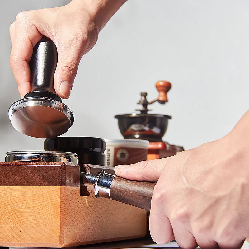 Person using a coffee tamp to compress coffee grounds in a portafilter with a coffee grinder in the background.