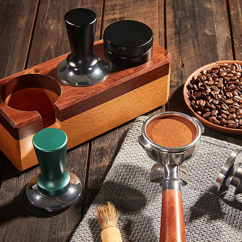 Coffee-making tools including a wooden tamper, green and silver tampers, and a bowl of coffee beans on a wooden surface.