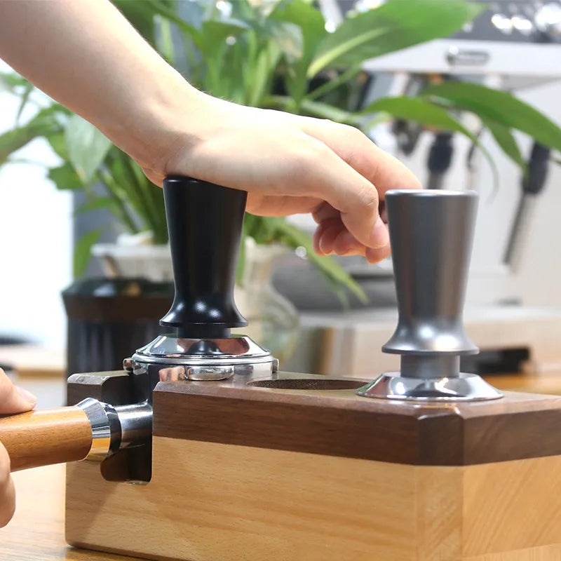 Hand using a coffee tamp to compress grounds in a wooden press with a blurred indoor background