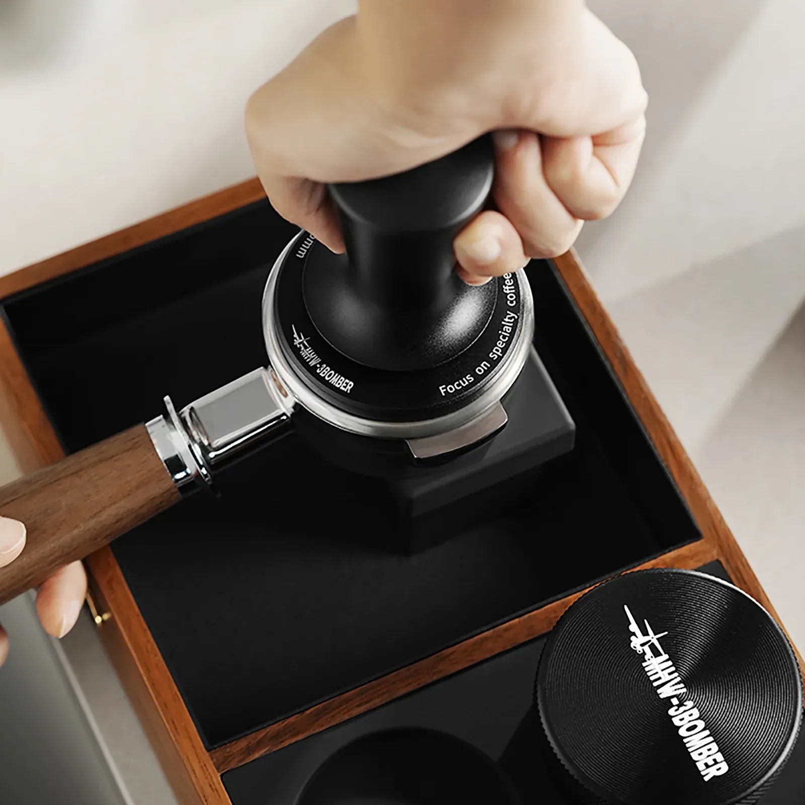 Hand using a coffee tamp with a wooden handle on a black tray, branded 'Hario'.