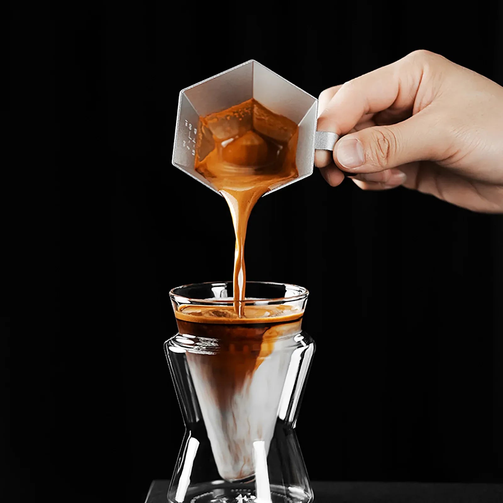 Hand pouring coffee from a white filter into a glass container on a black background