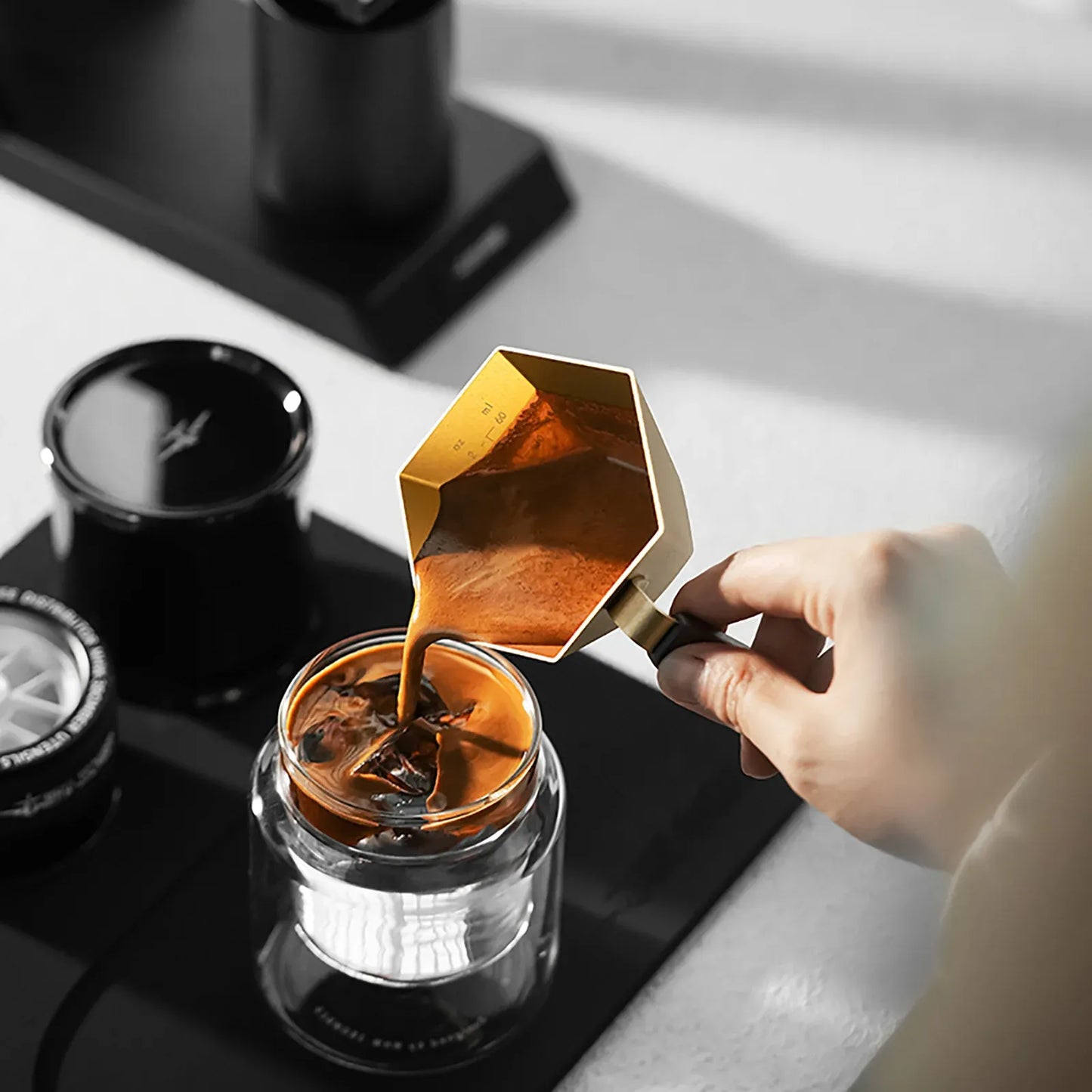 Hand pouring coffee from a gold Moka pot into a glass container on a kitchen counter.