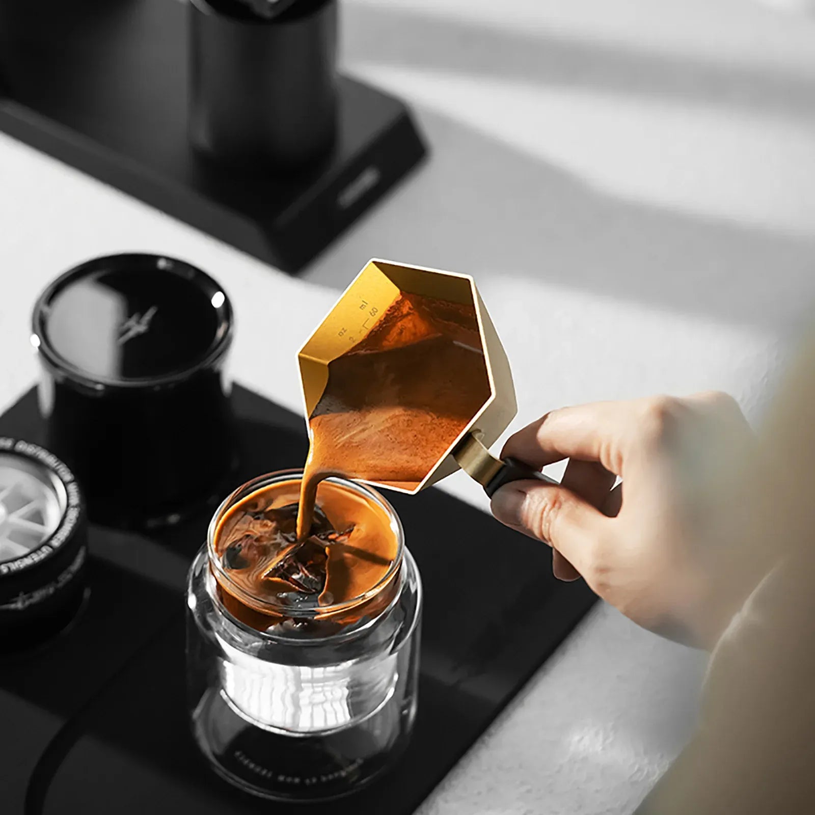 Hand pouring coffee from a gold Moka pot into a glass container on a kitchen counter.