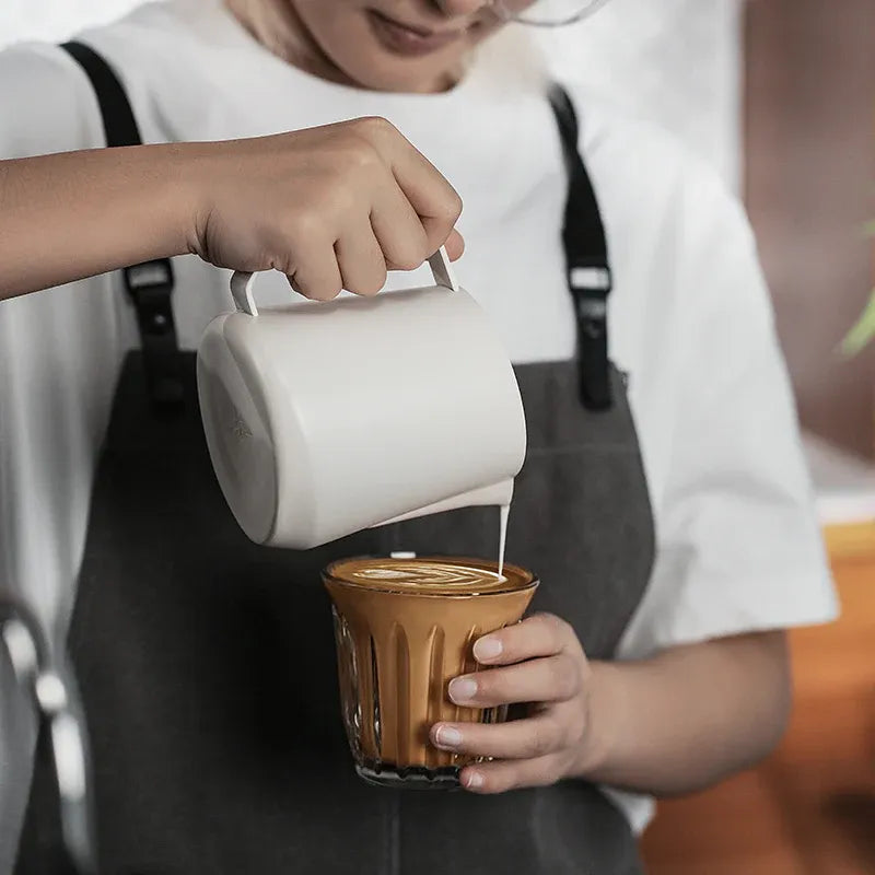 Person pouring milk from a white pitcher into a glass of coffee.