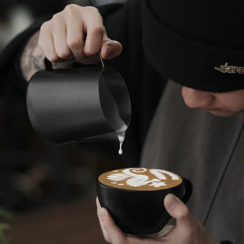 Person pouring milk into a black mug with latte art, wearing a black shirt with a logo.
