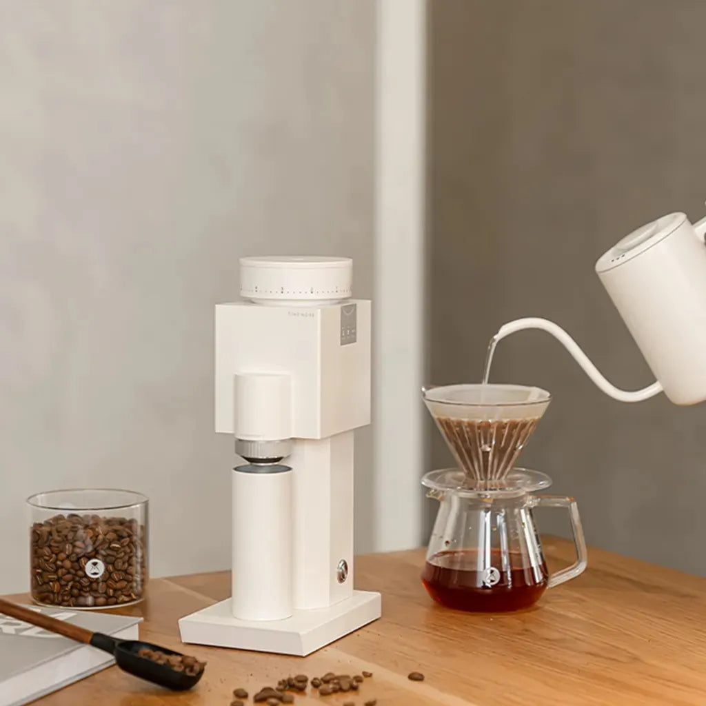 White coffee grinder and pour-over setup on a wooden surface with coffee beans and a glass container.