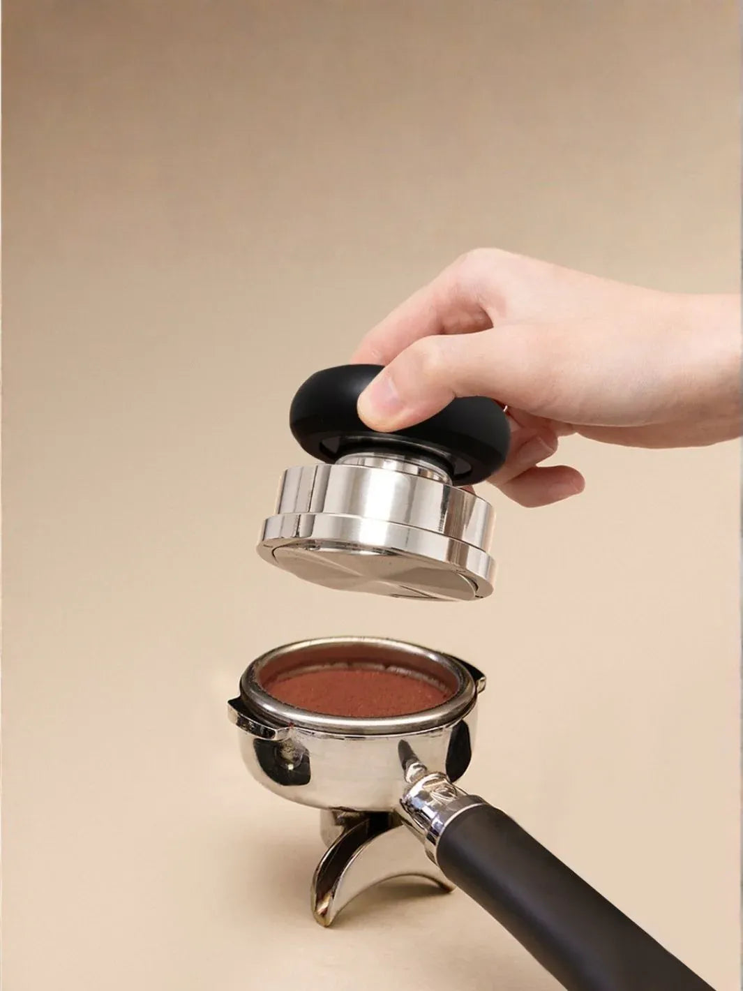 Hand using a coffee tamp to compress coffee grounds in a portafilter on a beige background