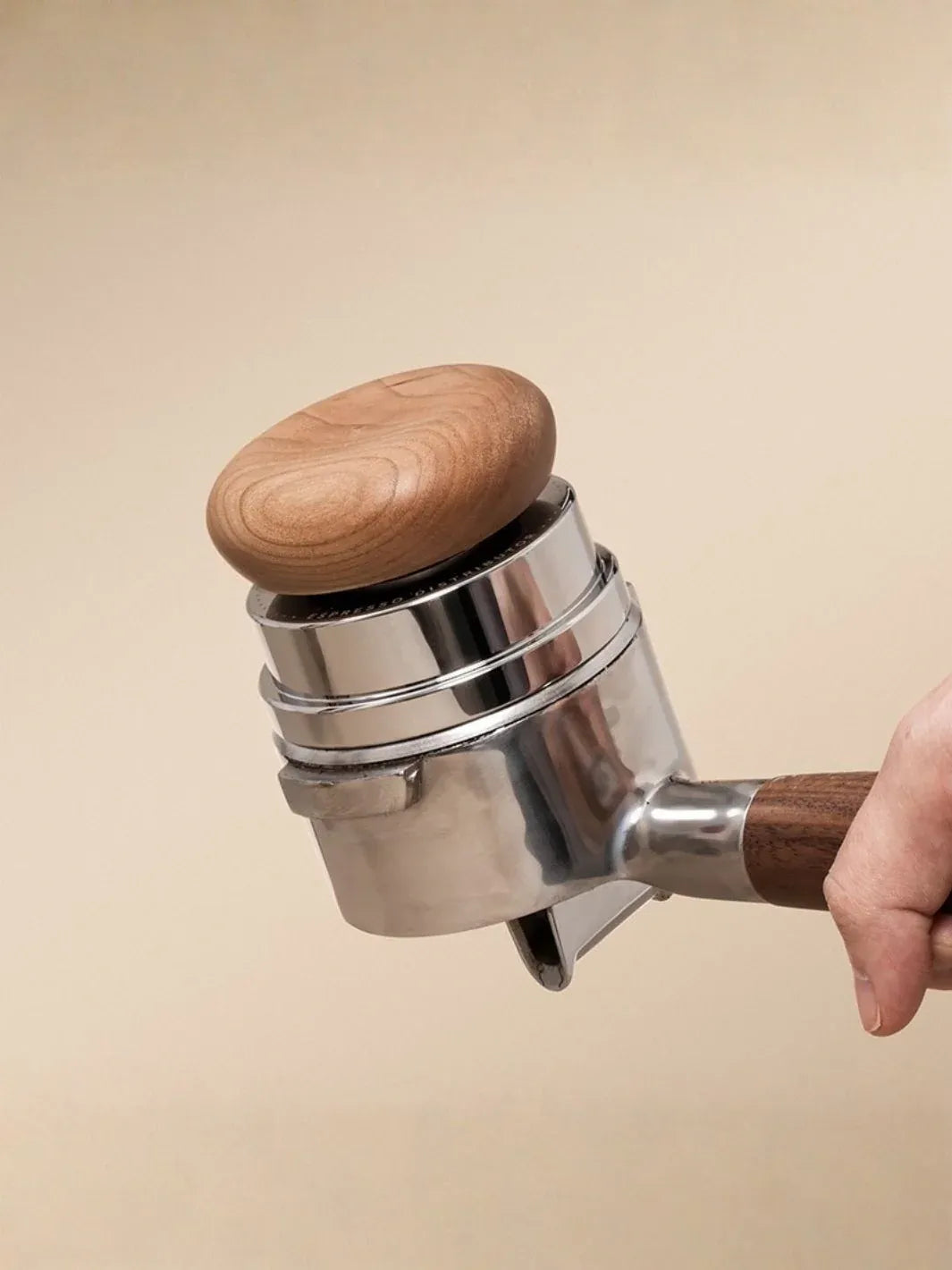 Hand holding a metal espresso tamper with wooden top and handle against a beige background