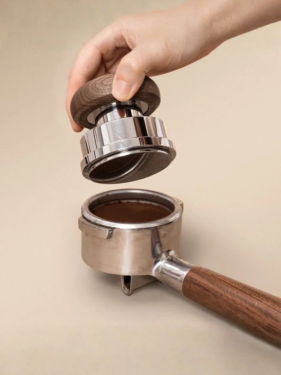 Hand using a coffee tamper to compress coffee grounds into a portafilter on a beige background