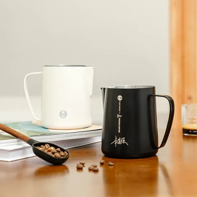 Black milk jug with white text on a wooden surface, next to a white mug and a small spoon with coffee beans.