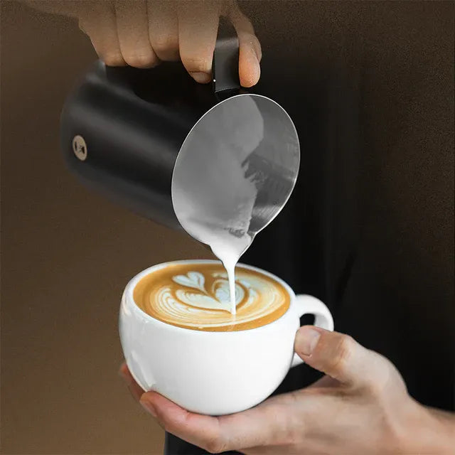 Person pouring milk from a pitcher into a cup of coffee with latte art.