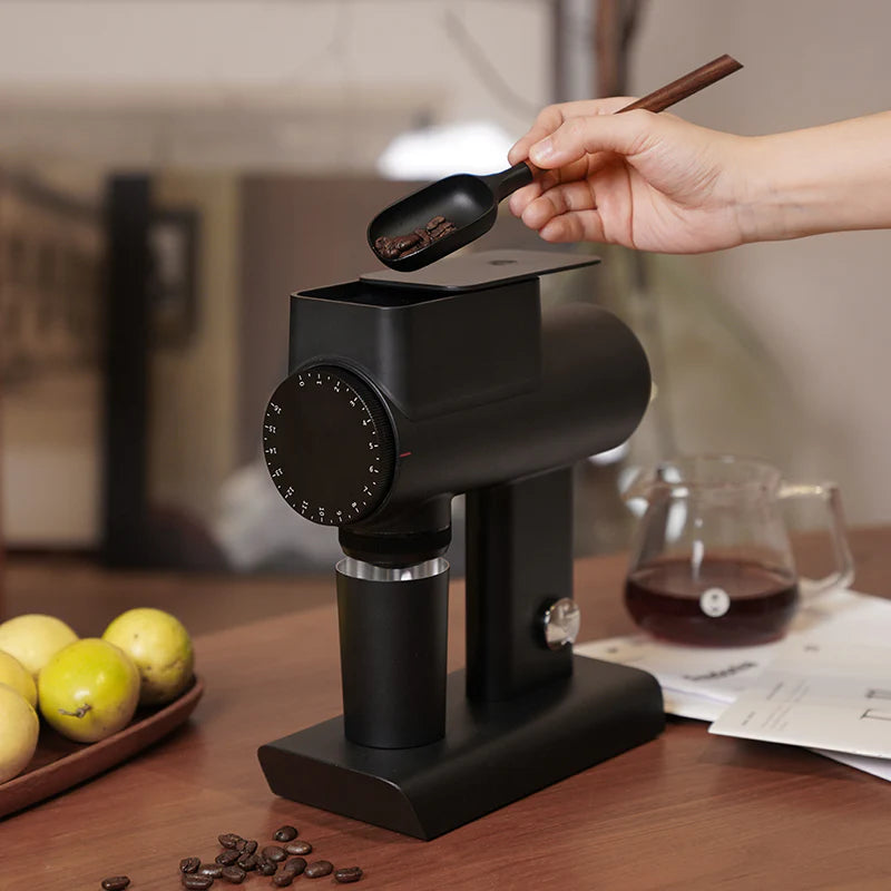 Black coffee grinder with a hand holding a scoop of ground coffee on a wooden table.