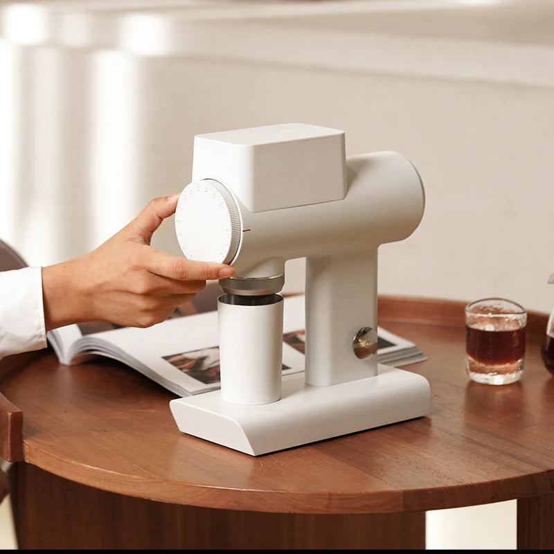 White coffee grinder on a wooden table with a glass of coffee and magazine in the background.
