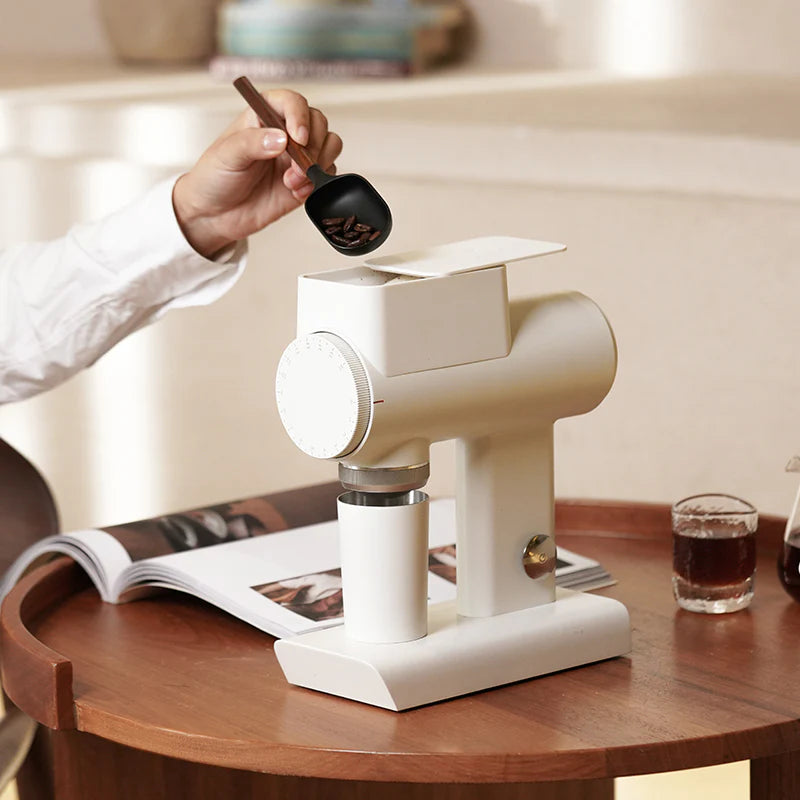 Person using a manual coffee grinder on a wooden table with a glass of coffee in the background.