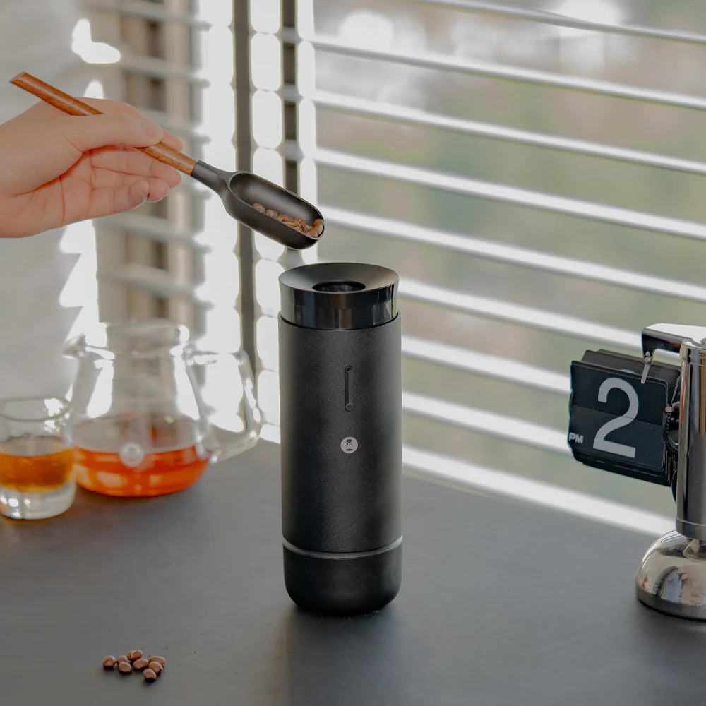 Black coffee grinder on a kitchen counter with a hand holding a scoop of coffee beans.